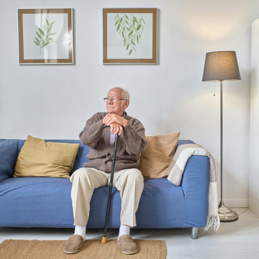 Verhinderungspflege - Heinrich-Gerold-Haus Ein älterer Mann sitzt auf einem blauen Sofa mit einem Stock, umgeben von Pflanzenbildern.