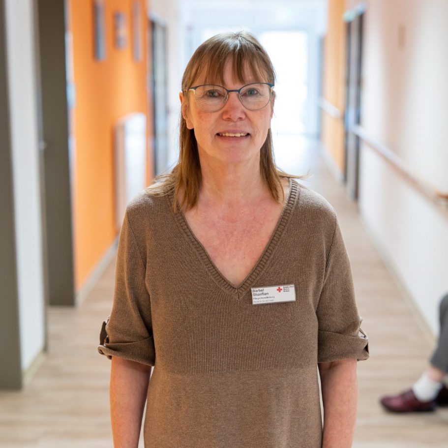 Bärbel Sharifian Pflegedienstleitung - Heinrich-Gerold-Haus Frau mit Brille und braunem Oberteil lächelt in einem Flur mit orangefarbenen Wänden.